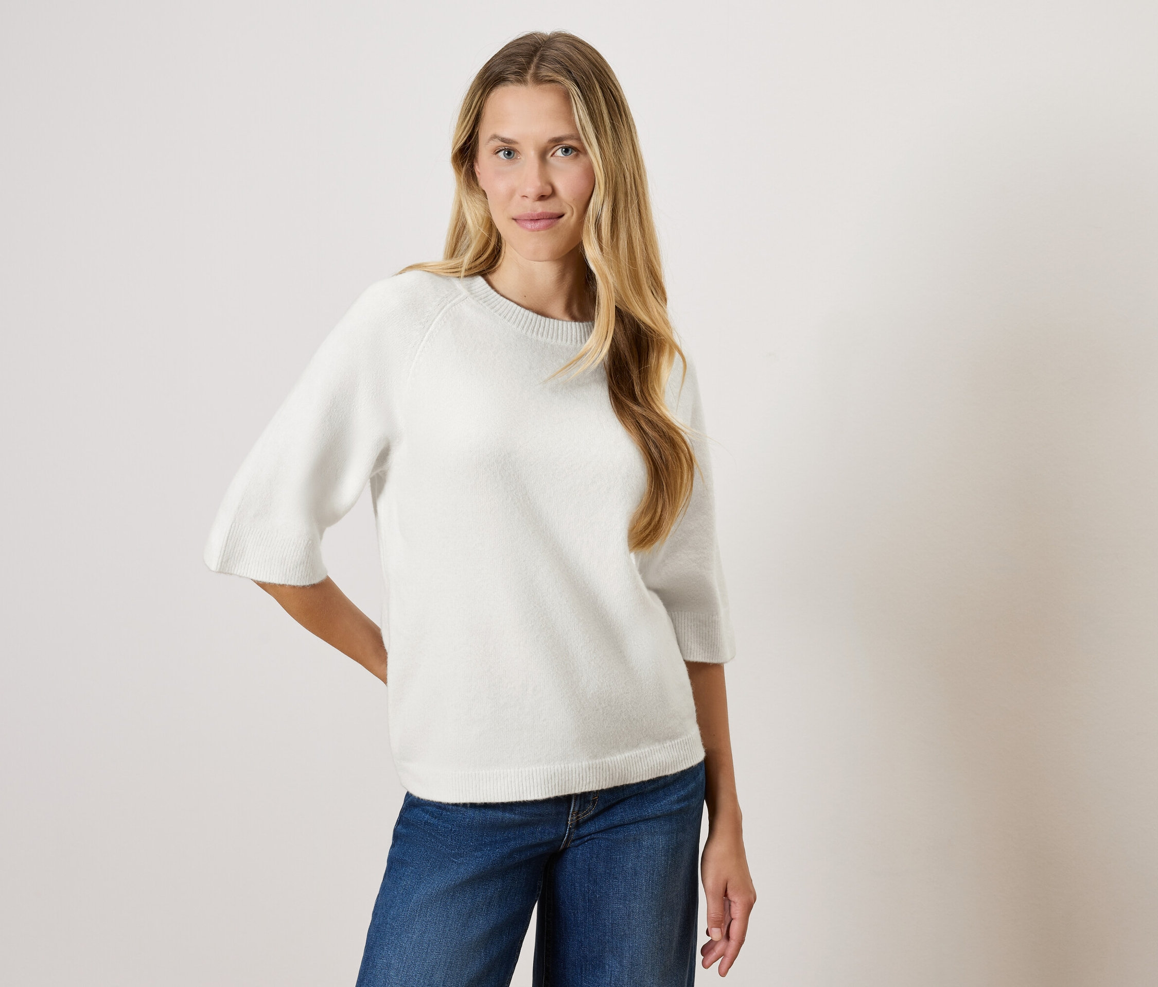 Žena s dlhými blond vlasmi pózuje v bielom svetri s trojštvrťovými rukávmi a modrých džínsoch.