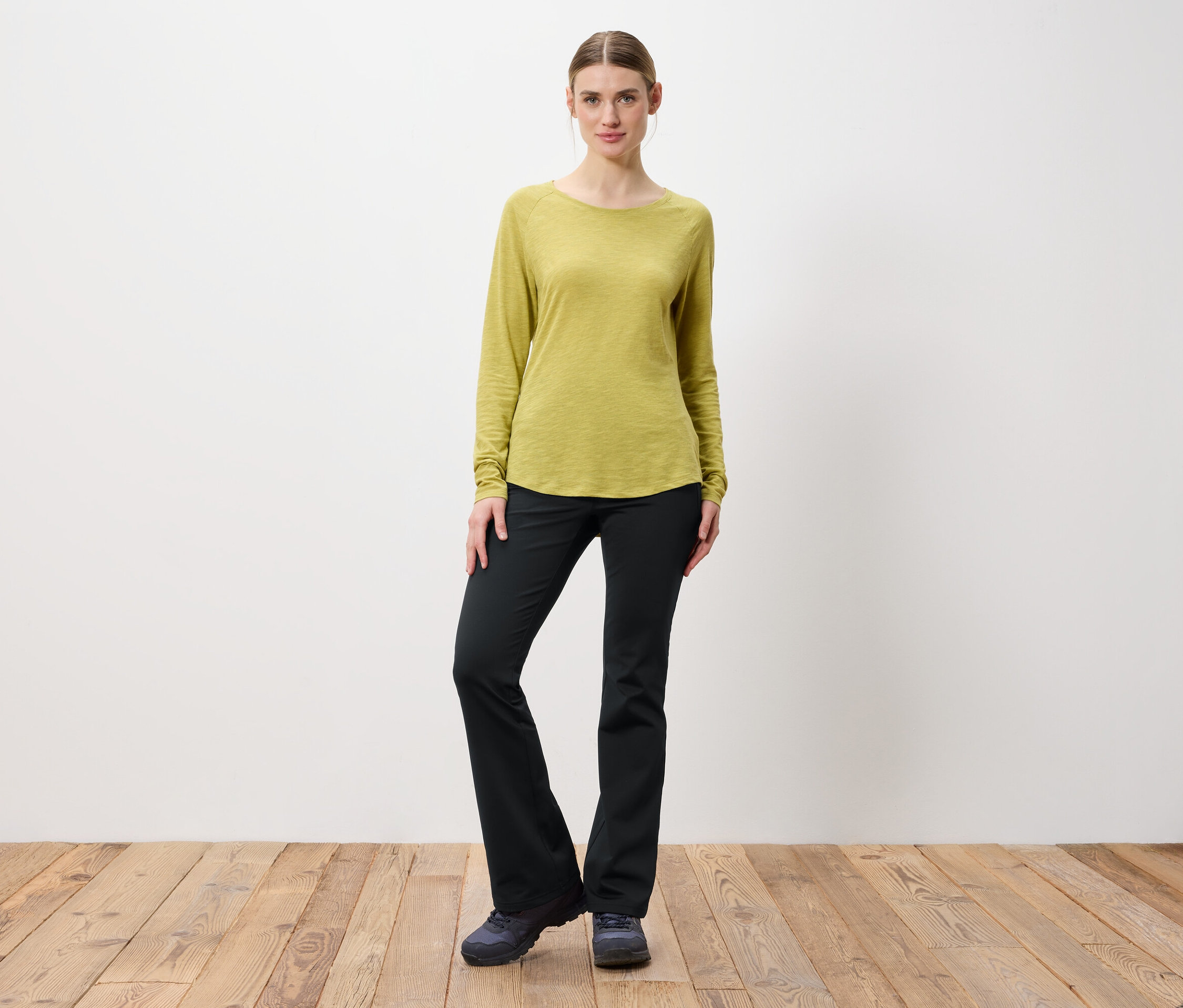 Žena pózuje v svetlozelenom tričku s dlhým rukávom a čiernych nohaviciach.