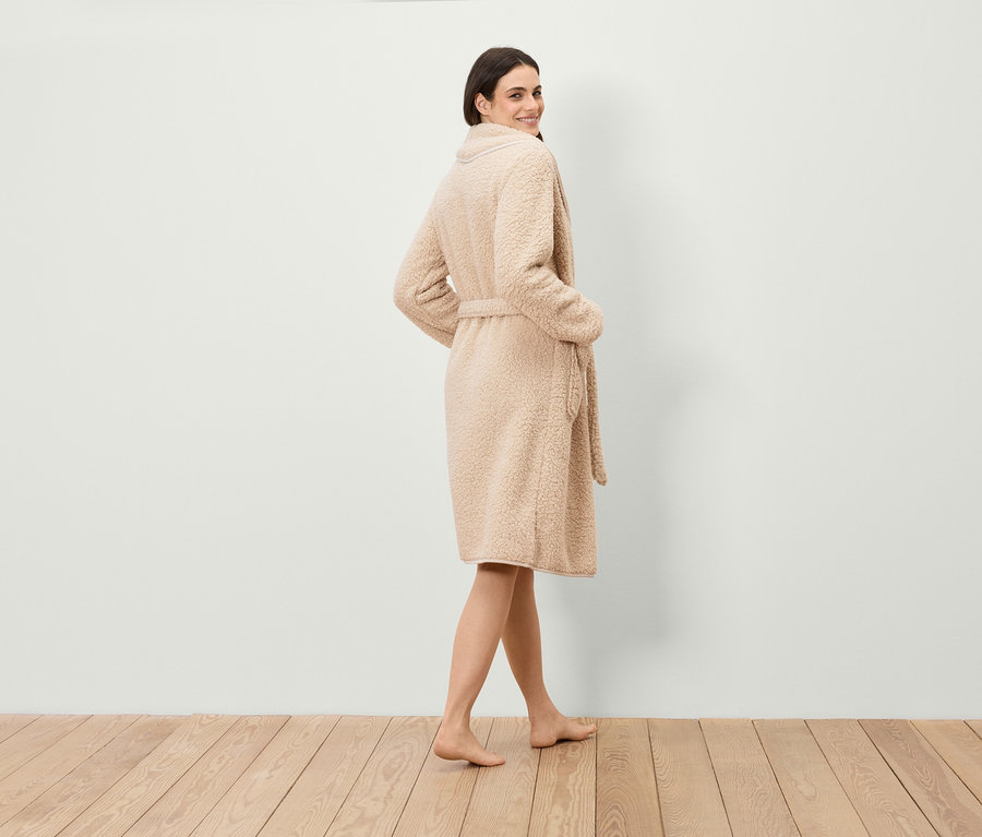 Žena sa pozerá cez plece, má na sebe župan z umelej kožušiny s opaskom a stojí na drevenej podlahe.