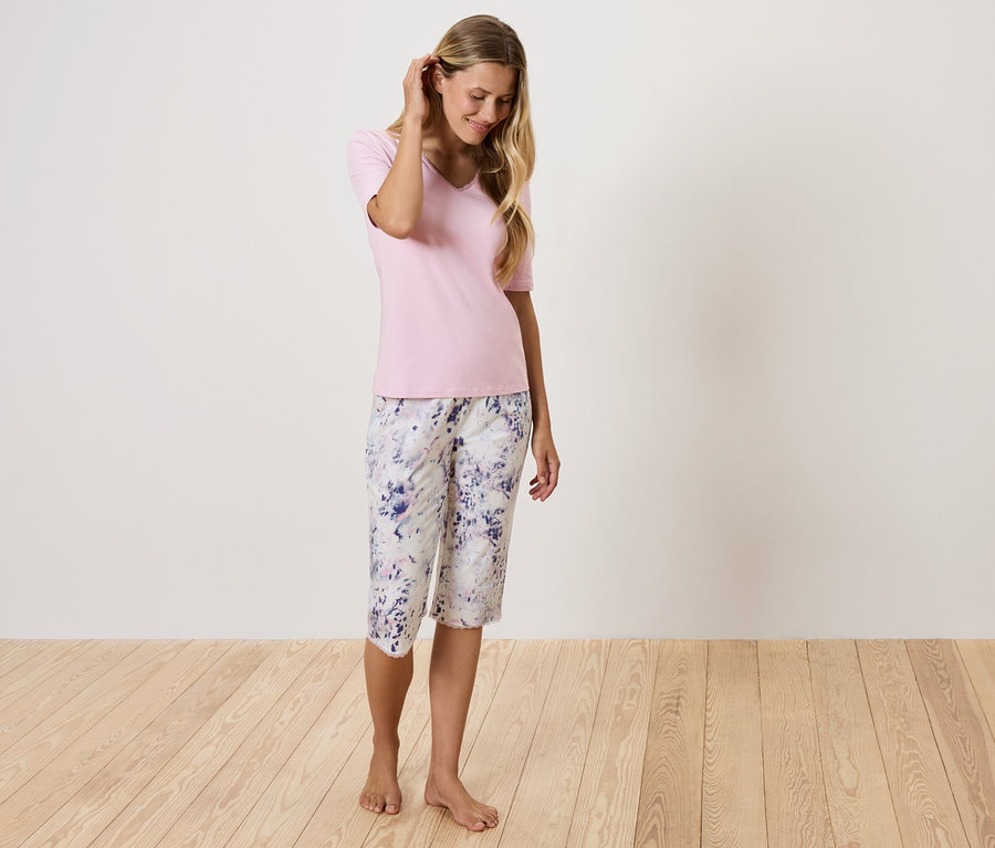 Žena v ružovom tričku a kvetinových nohaviciach stojí na drevenej podlahe.
