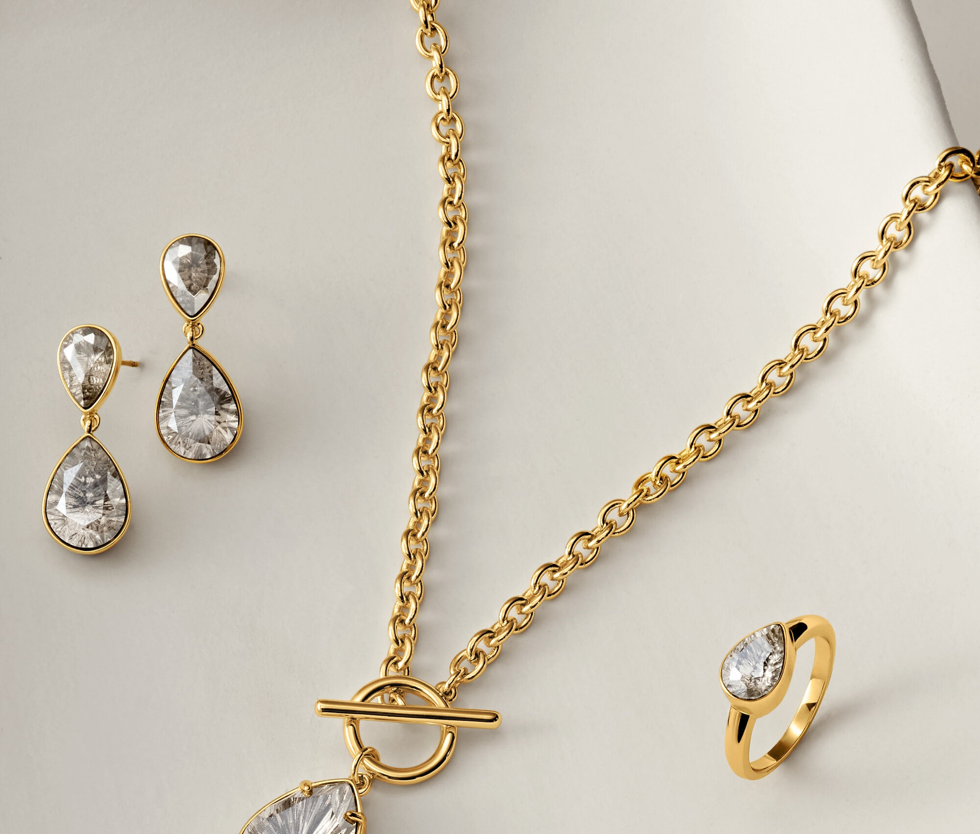 Zlatý náhrdelník, náušnice a prsteň s čírymi kameňmi v tvare slzy na bielom pozadí.