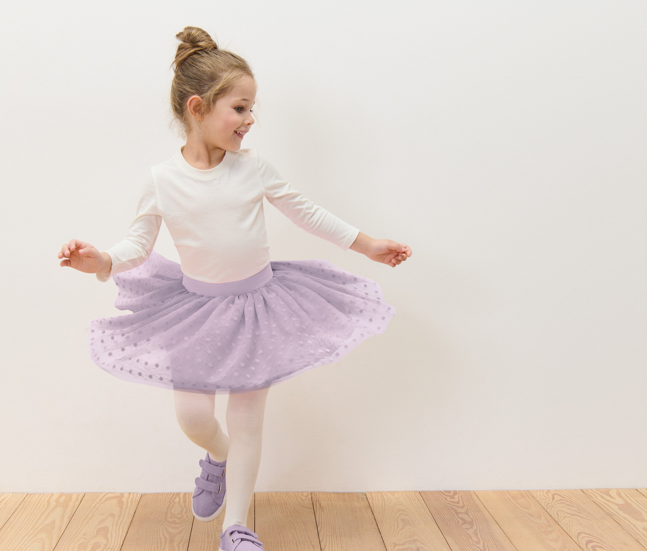 Dievča s drdolom tancuje v fialovej tylovej sukni s bielymi bodkami, bielom tričku s dlhým rukávom, pančuchách a fialových topánkach pred bielou stenou.