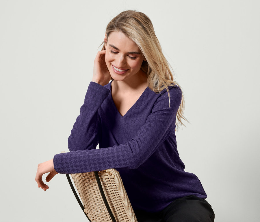 Blondínka sedí na prútenej stoličke, má na sebe fialové tričko s dlhým rukávom a usmieva sa.
