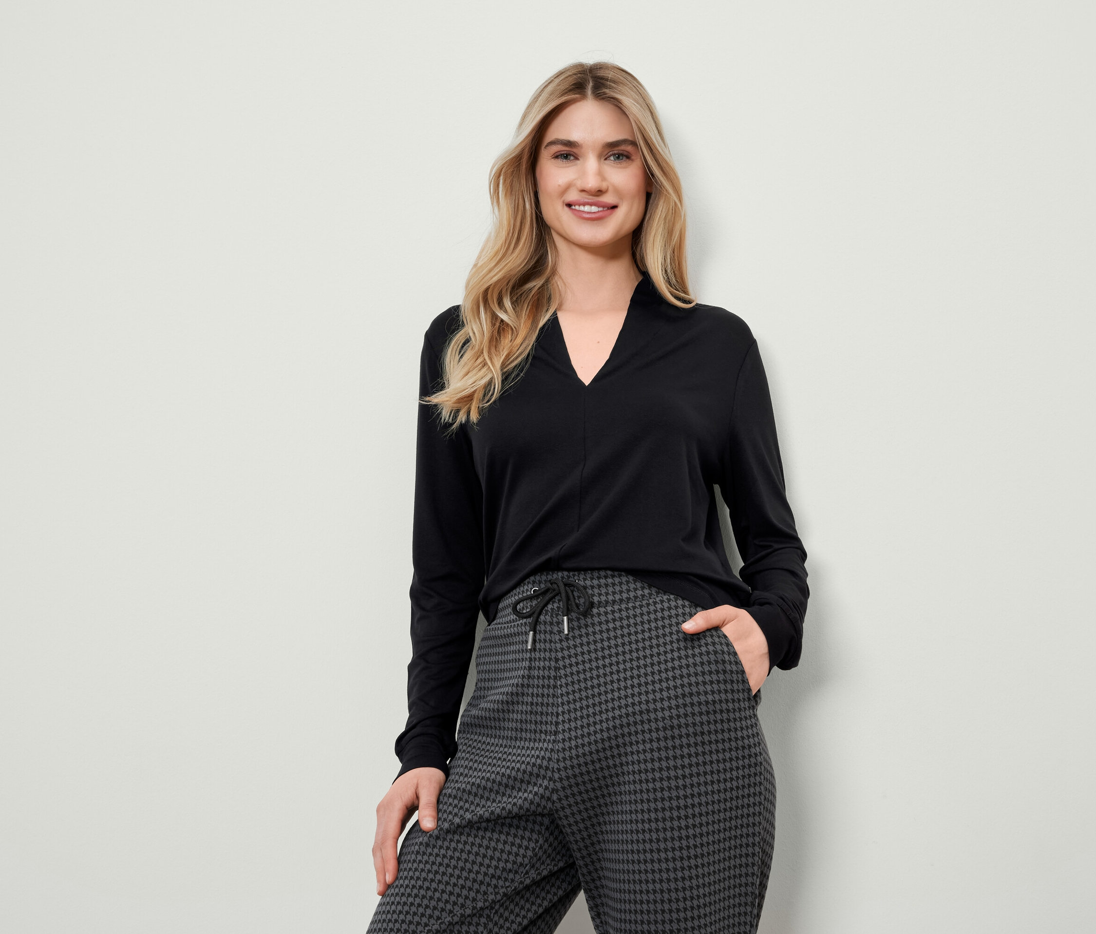 Blondínka v čiernom tope s dlhým rukávom a sivých nohaviciach so vzorom kohútej stopy pózuje.