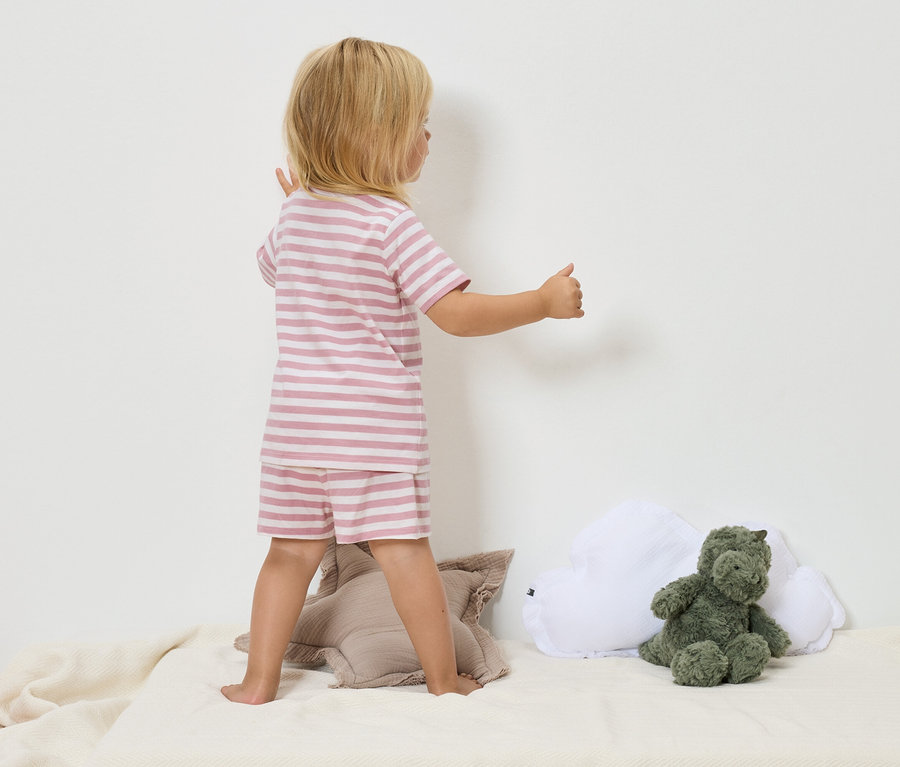 Dieťa s blond vlasmi v ružovo-bielom pásikavom pyžame stojí na béžovej deke vedľa zelenej plyšovej hračky dinosaura a bieleho obláčikového vankúša.