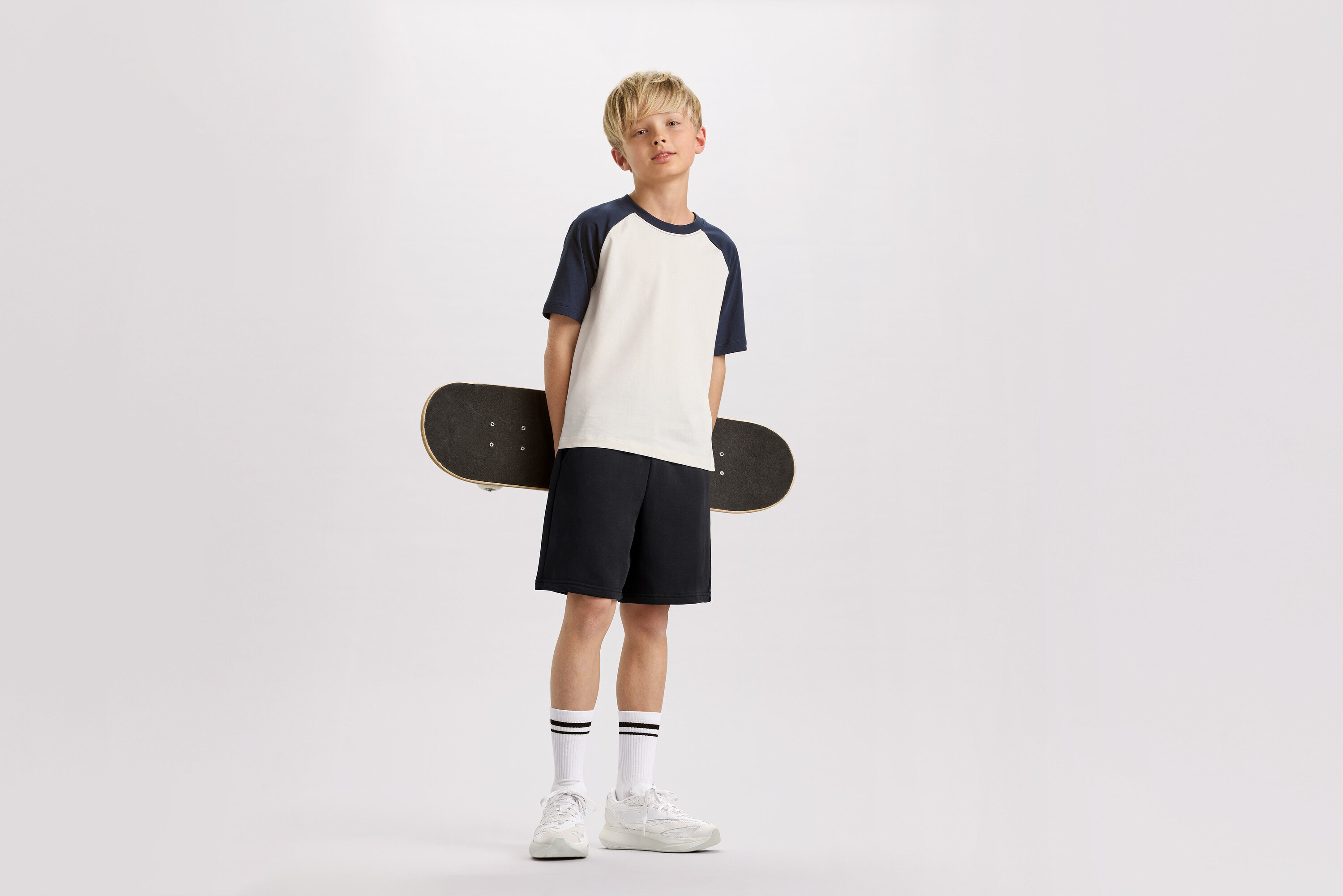 Blond chlapec drží za chrbtom skateboard, má na sebe tričko s tmavomodrými rukávmi, šortky a tenisky.
