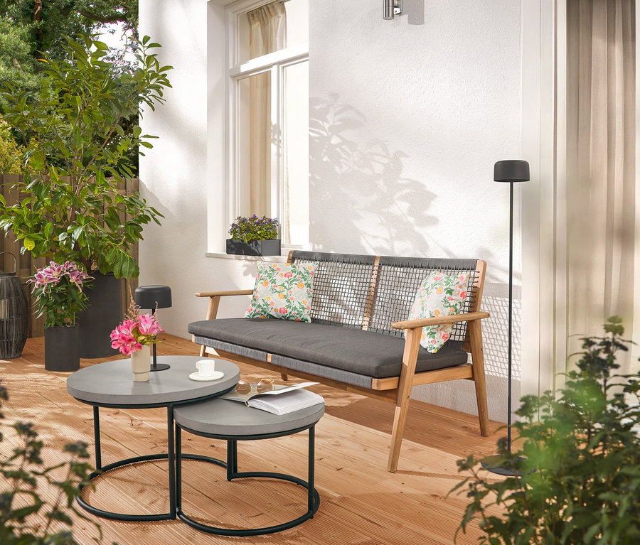 Na terase sa nachádza pohovka s vankúšmi, dva sivé stolíky, stojaca lampa a rastliny.