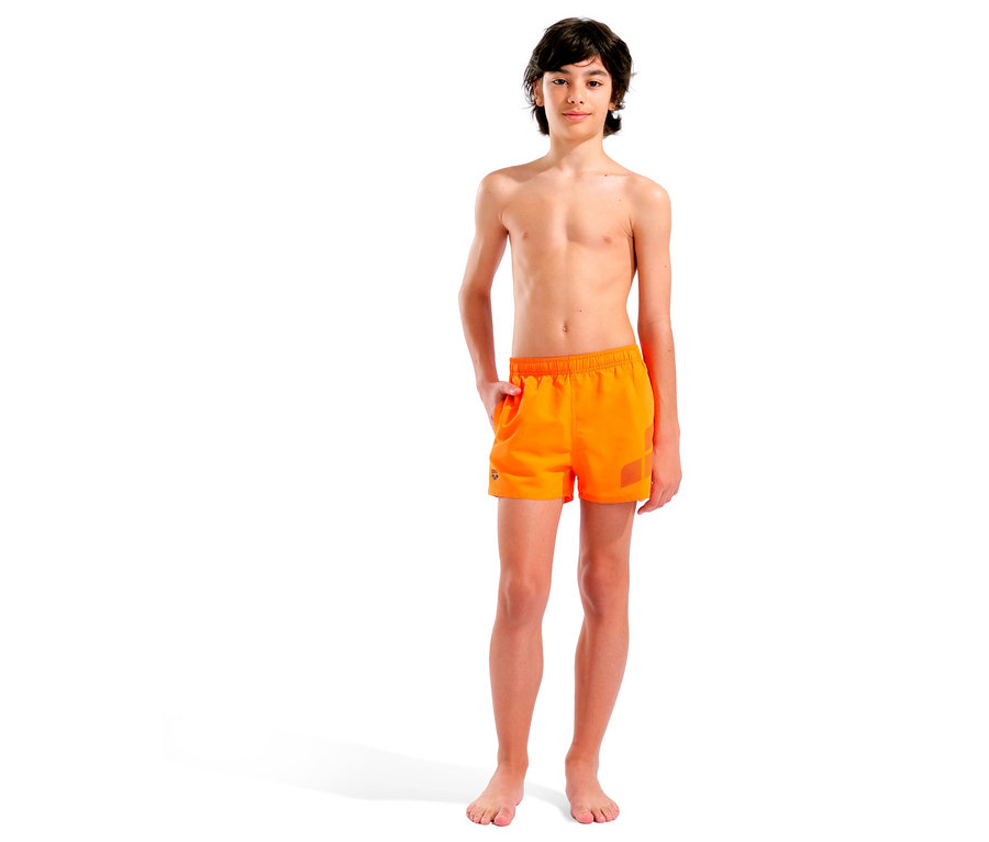 Chlapec stojí bosý na bielom pozadí a má na sebe oranžové plavky.