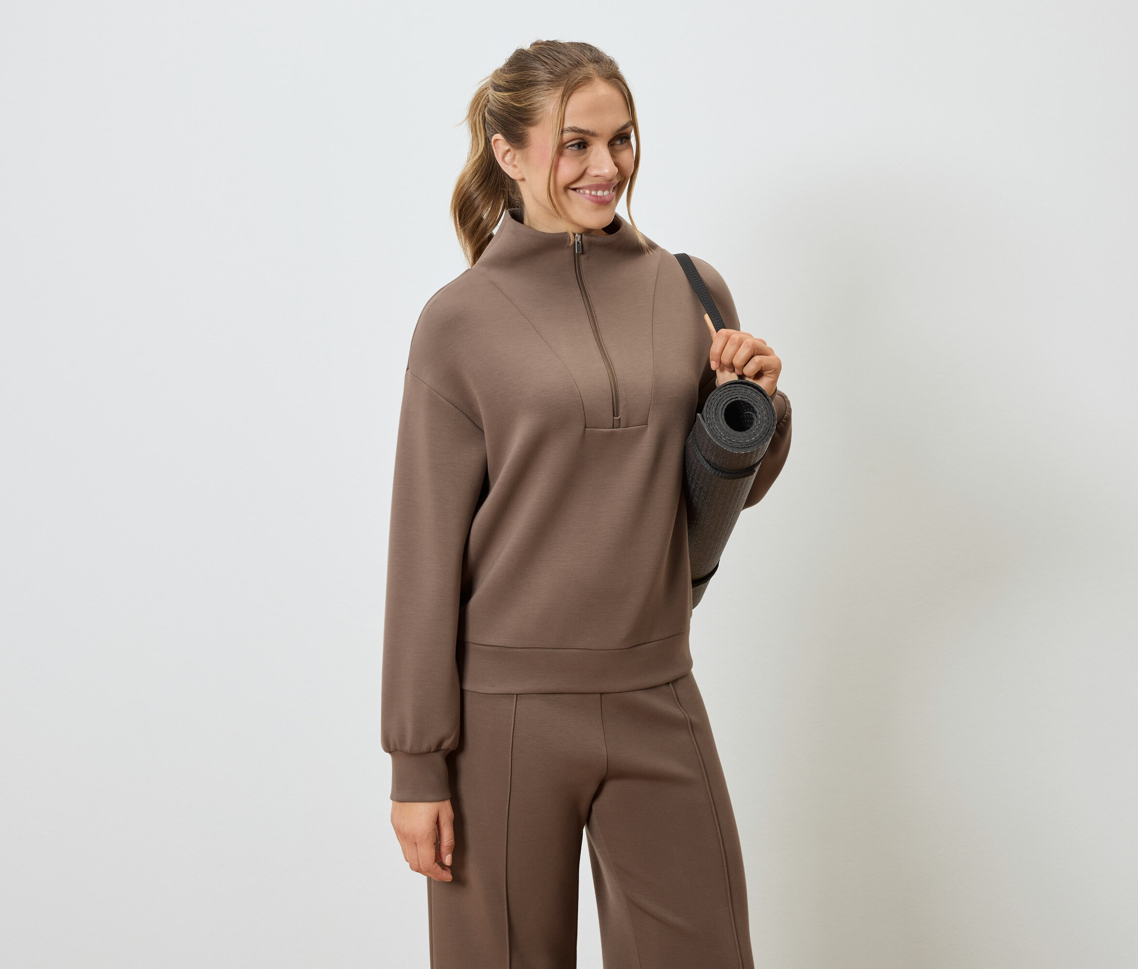 Žena v hnedej teplákovej súprave s podložkou na jogu prehodenou cez rameno.