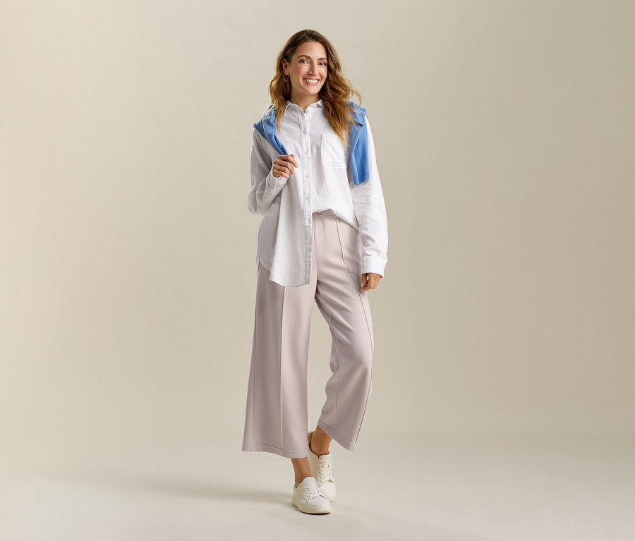 Usmievavá sa žena v bielej, dlhej ľanovej blúzke a pieskových nohaviciiach culotte zo zmesi modalu. Na ramenách má modrý sveter.