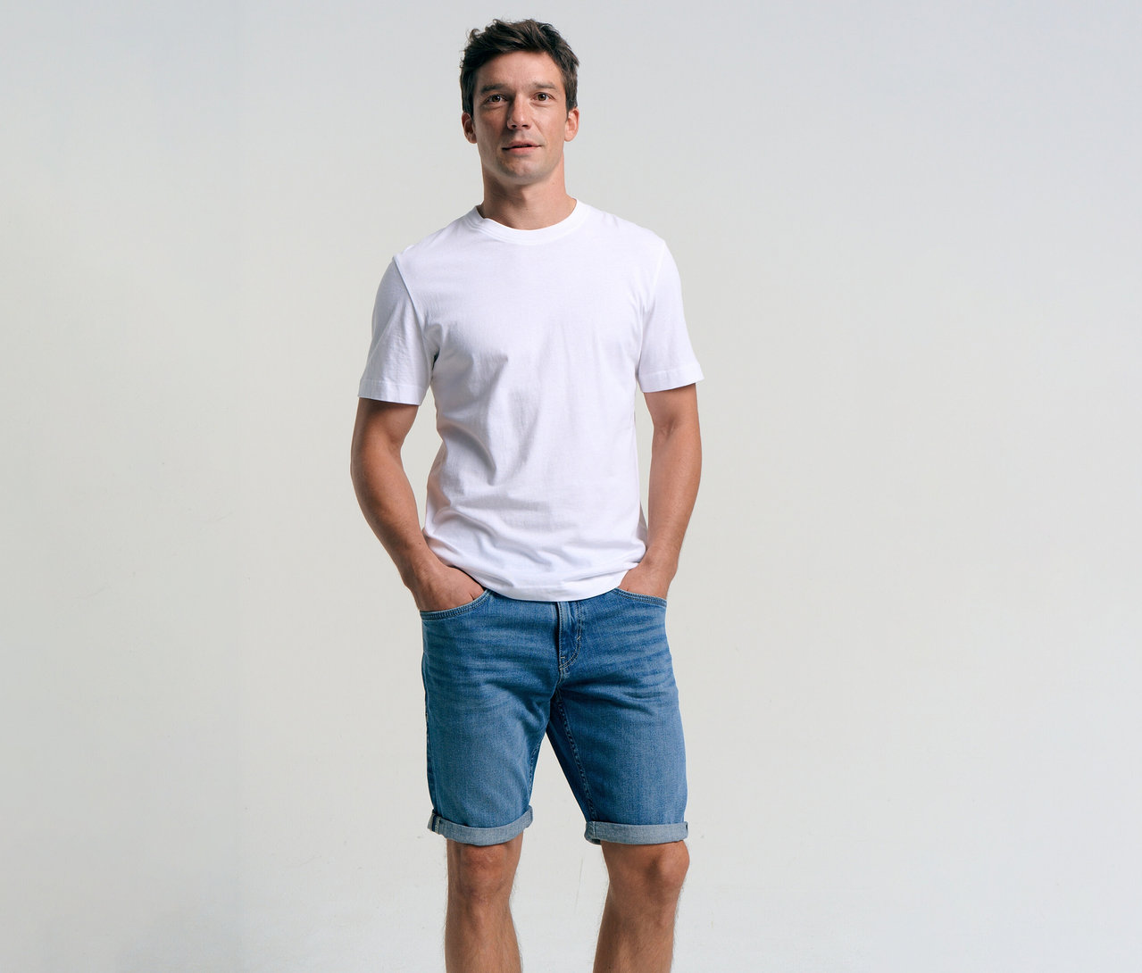 Muž v bielom tričku a džínsových šortkách Tom Tailor TTJOSH REGULAR SLIM, mid stone bright blue denim, s rukami vo vreckách.