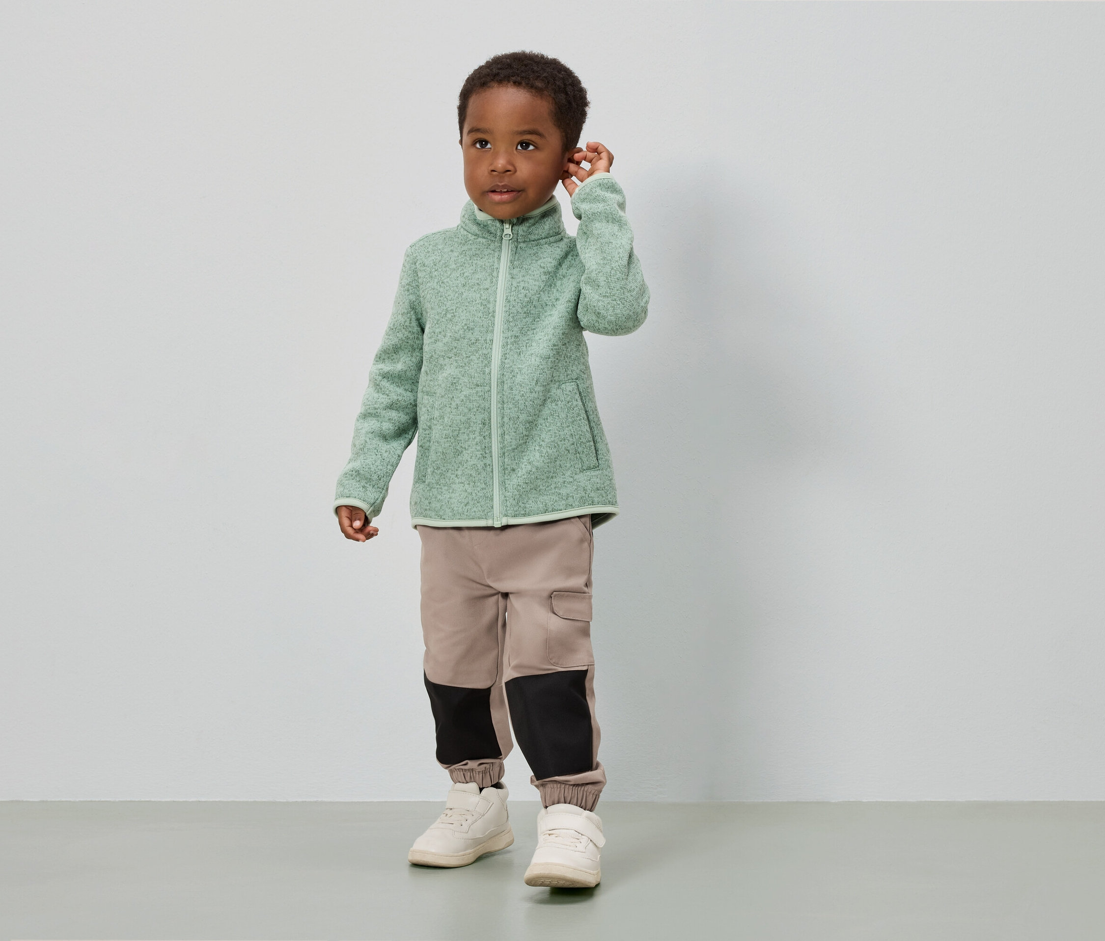 Chlapec stojí pred bielou stenou a má na sebe svetlozelenú fleecovú bundu, béžové nohavice s čiernymi vložkami a biele topánky.