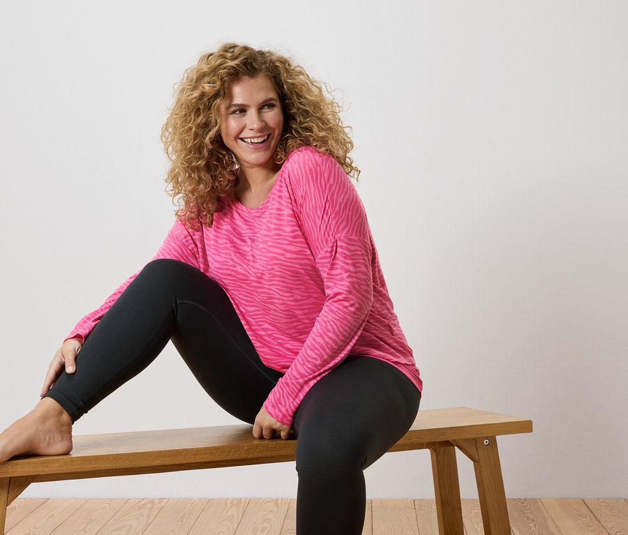 Žena s kučeravými vlasmi sedí naboso na drevenej lavičke, má na sebe ružové tričko s dlhým rukávom so vzorom zebry a čierne legíny.