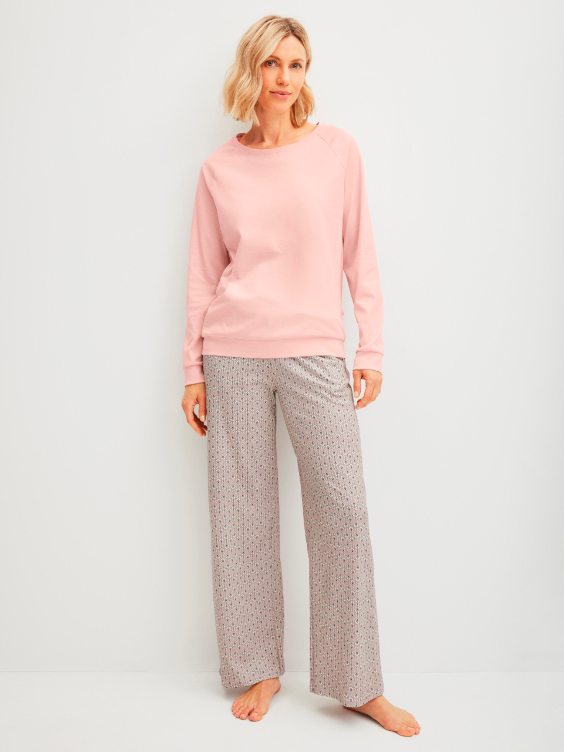 Žena stojí naboso v ružovom svetri a vzorovaných nohaviciach pred bielou stenou.