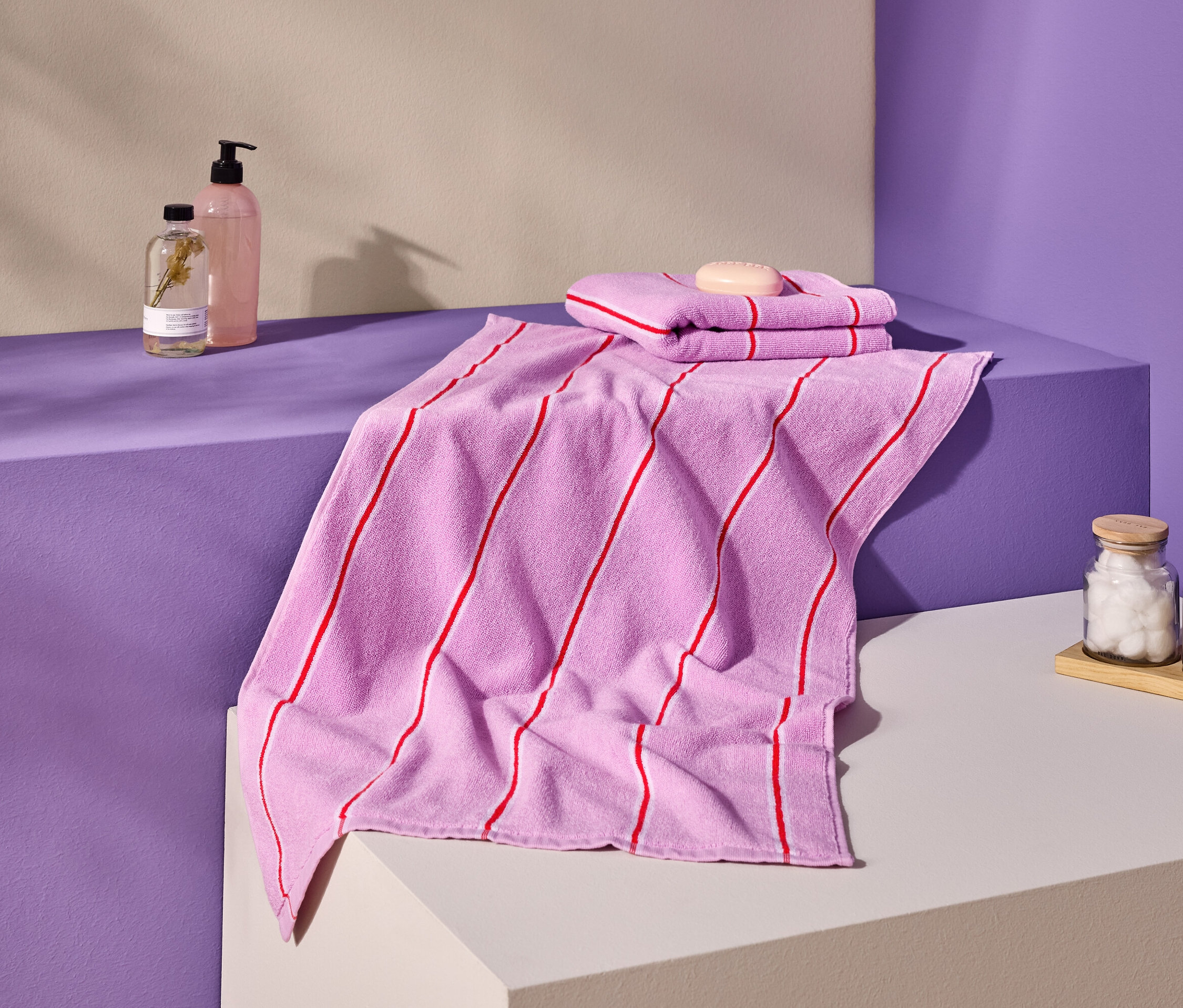 Snímka ružových uterákov s červenými pruhmi, mydla, fliaš a nádob na fialovom a krémovom povrchu.