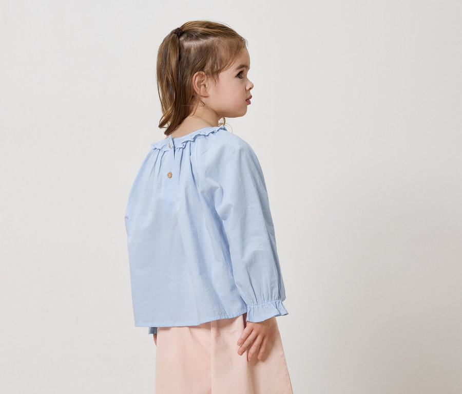 Dievča zozadu zboku s modrou blúzkou s volánmi a ružovými nohavicami na bielom pozadí.