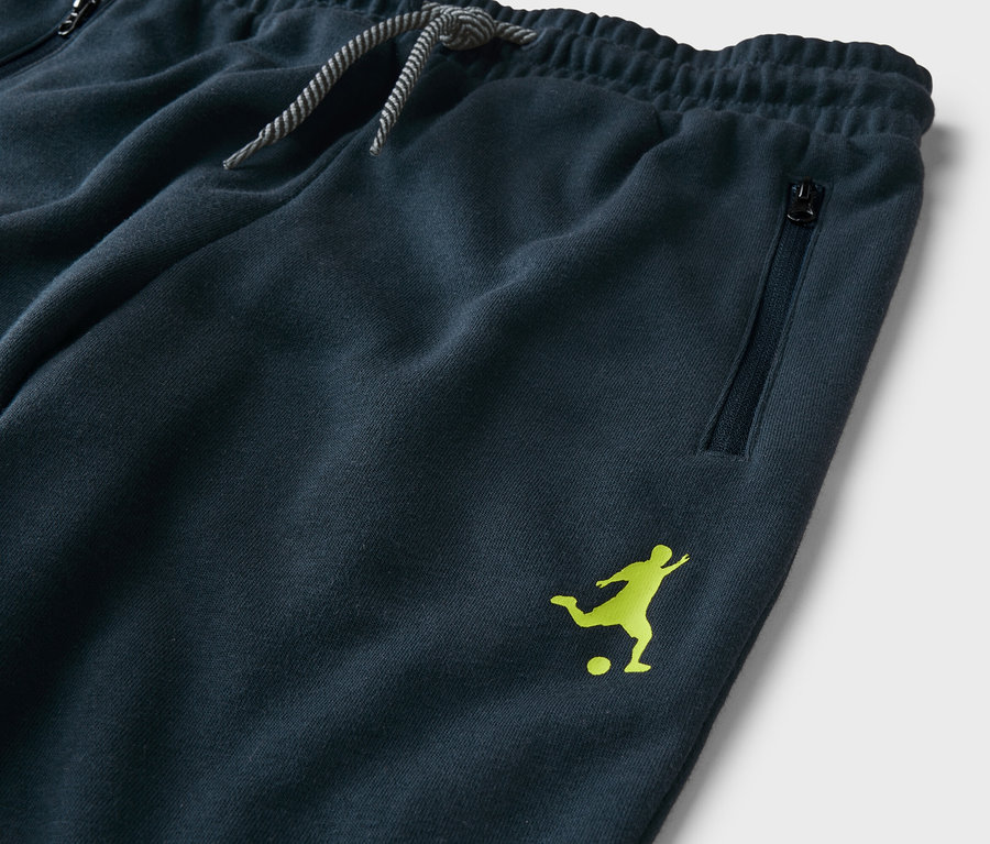 Detailný záber na námornícke modré tepláky so šnúrkou, vreckom na zips a svetlozeleným logom futbalistu.
