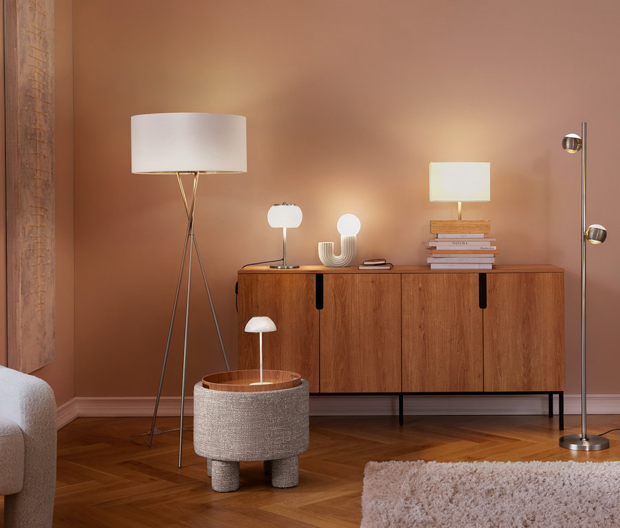 Obývacia izba s viacerými zapnutými lampami. Biela stolová LED lampa s akumulátorom stojí na okrúhlom príručnom stolíku, vedľa kresla a drevenej skrinky s ďalšími lampami.