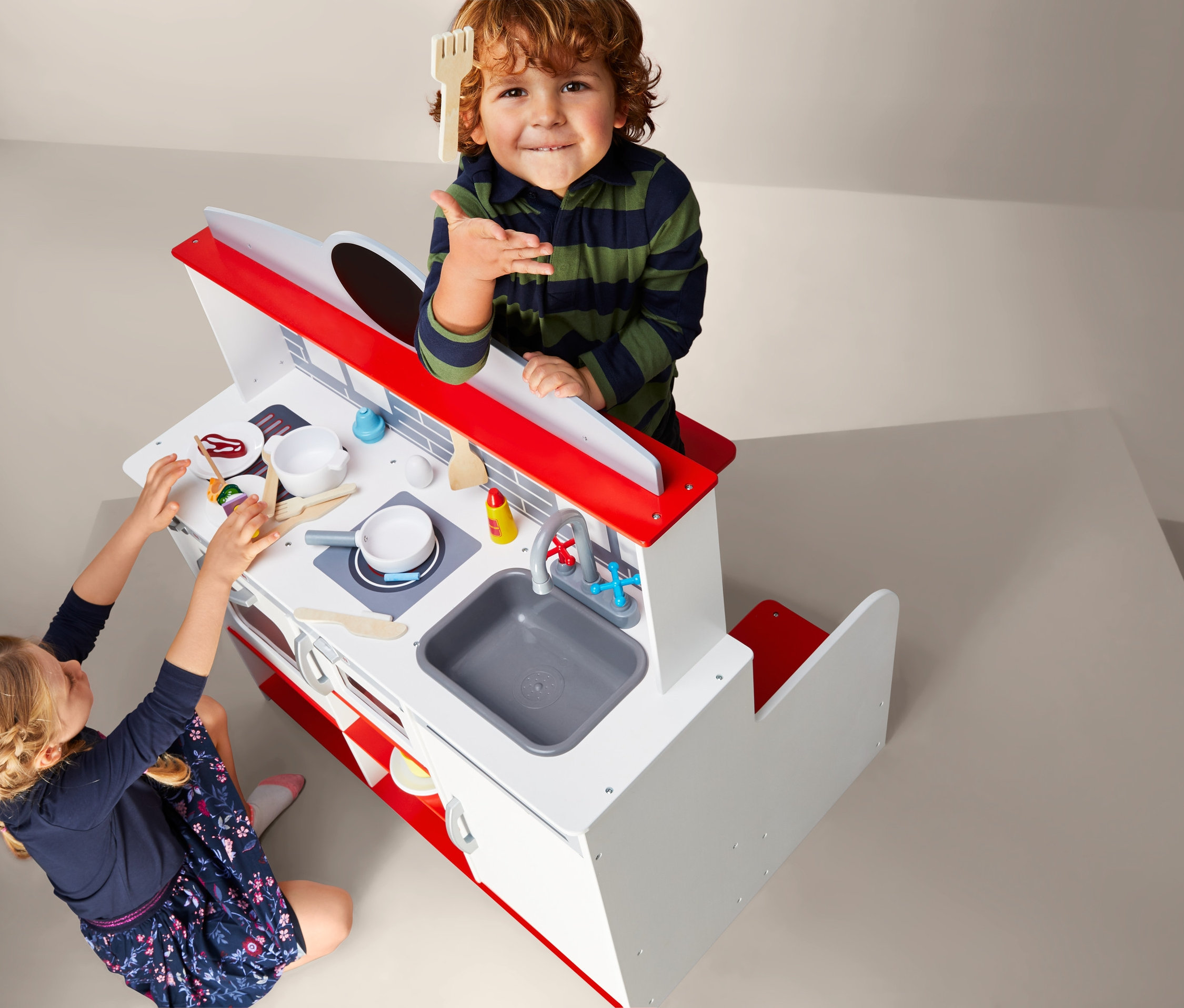 Deti sa hrajú s 2 v 1 reštauračným kútikom s kuchynkou. Chlapec drží drevenú vidličku, zatiaľ čo dievča sa hrá s jedlom na kuchynskej linke.