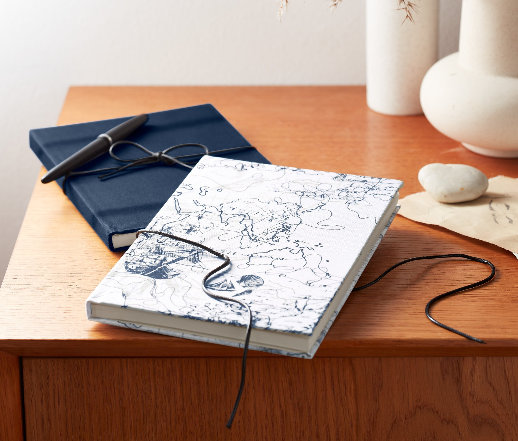 Dva zápisníky ležia na drevenej komode. Modrý zápisník s perom a biely s mapou.