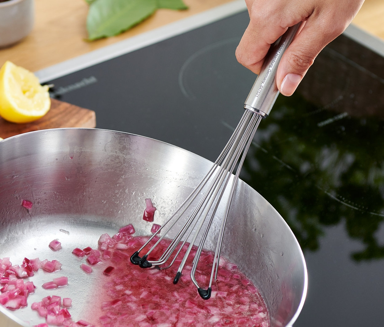 Ruka mieša profesionálnou metličkou nakrájanú červenú cibuľu v hrnci na sporáku.