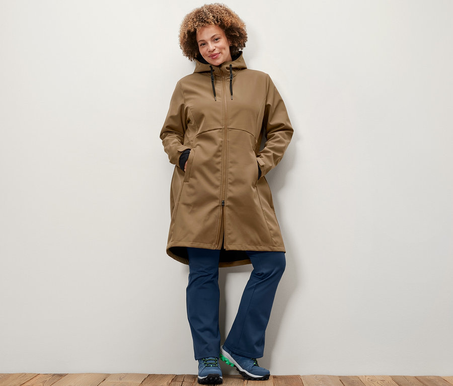 Žena pózuje v softshellovom kabáte nugátovej farby a rozšírených termonohaviciach.