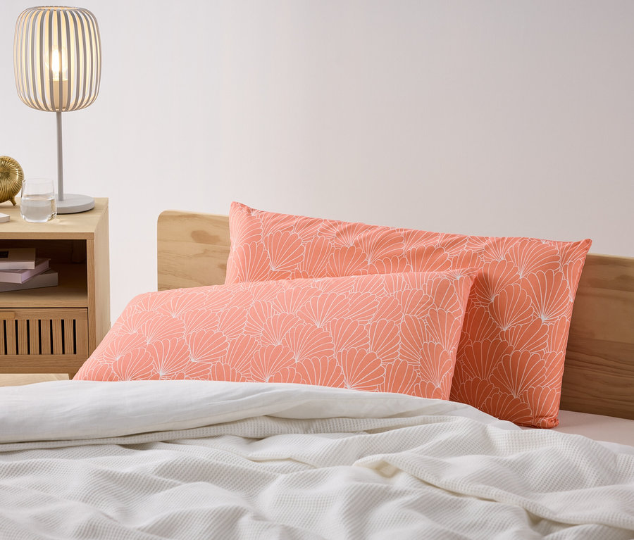 Dve koralové obliečky na vankúše z mikrovlákna s bielym kvetinovým vzorom ležia na posteli. Nočný stolík s lampou stojí vedľa postele.