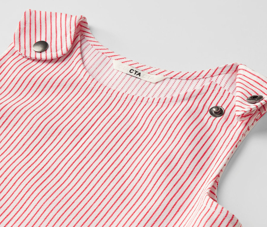 Detail detských tkaných pinaforových šiat s červenými prúžkami a kovovými gombíkmi.