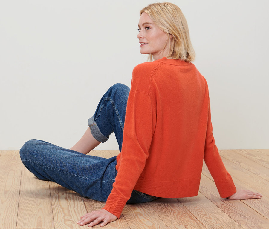 Blondínka sedí na drevenej podlahe a má na sebe oranžový pulóver z jemnej pleteniny s kašmírom a džínsy.