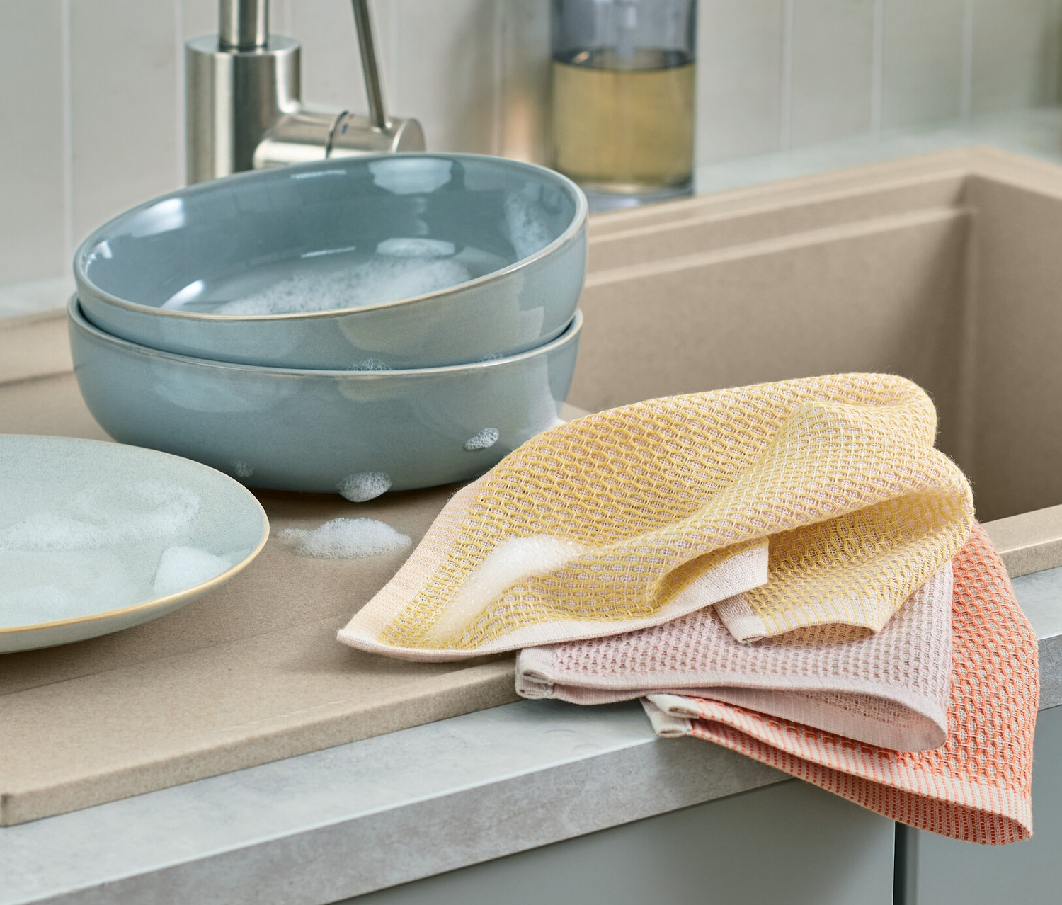 Dve modré misky s mydlovou vodou a jeden prázdny tanier ležia vedľa seba na odkvapkávači. Vedľa nich ležia 3 handričky na riad.