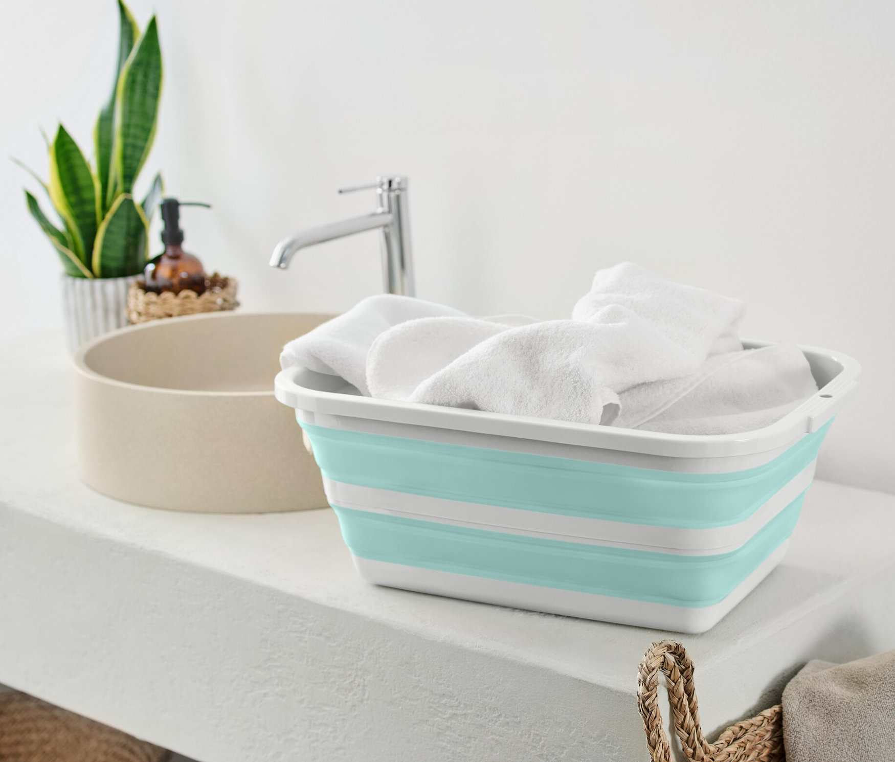 Skladací kôš na bielizeň plný bielych uterákov stojí na bielej poličke vedľa umývadla.