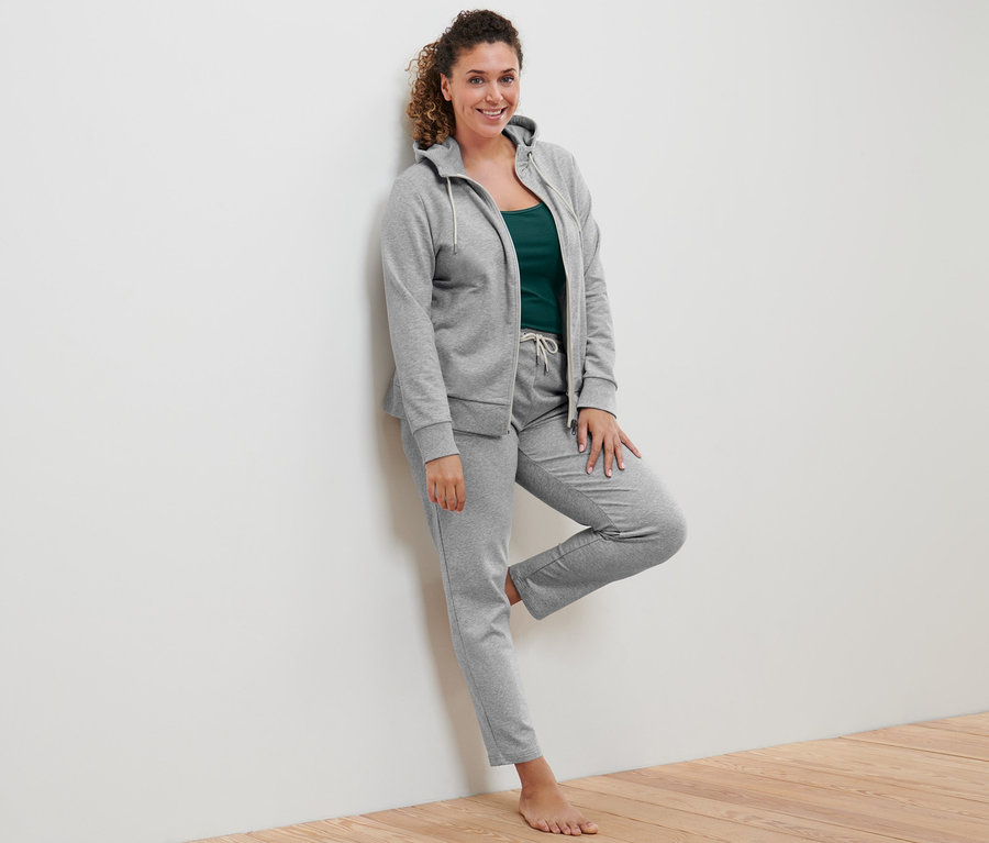 Žena opretá o stenu má na sebe sivú mikinu, zelené a sivobiele tielka (2 ks) a sivé tepláky.