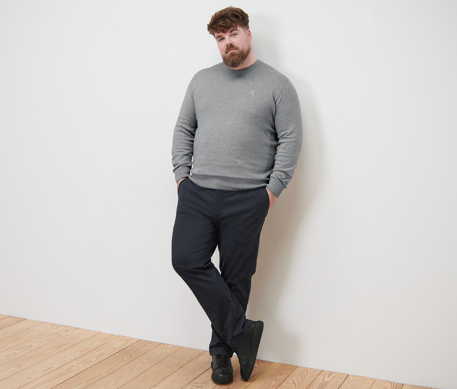 Muž sa opiera o stenu a má na sebe sivý pulóver s okrúhlym výstrihom a námornícke modré chino nohavice.