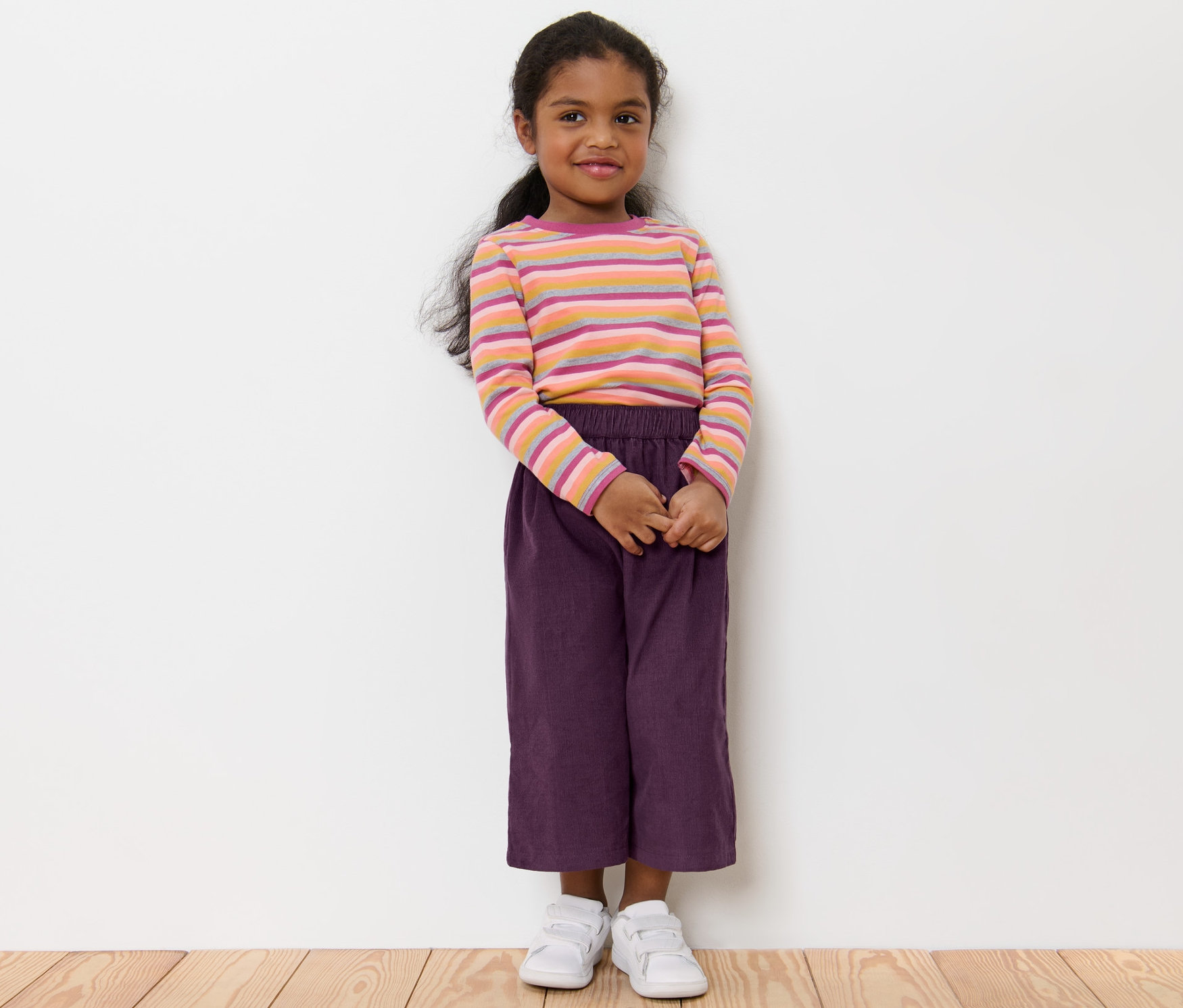 Dievča stojí a má na sebe detské nohavice typu culotte, pruhované tričko a biele topánky.