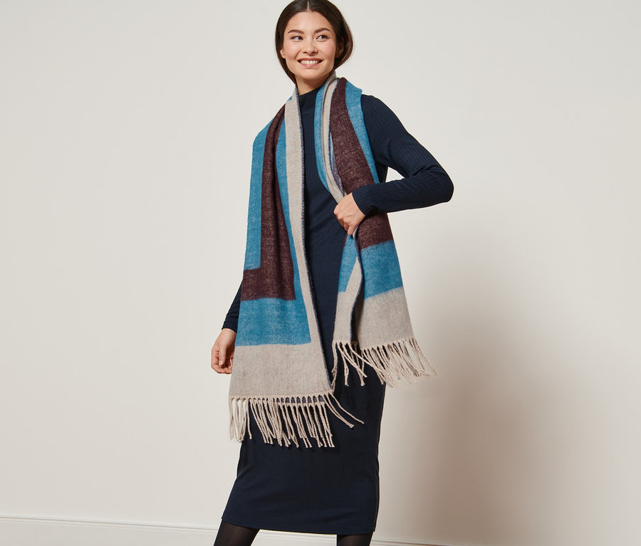 Žena pózuje s tkanou šálou s geometrickým vzorom v odtieňoch modrej, hnedej a béžovej.