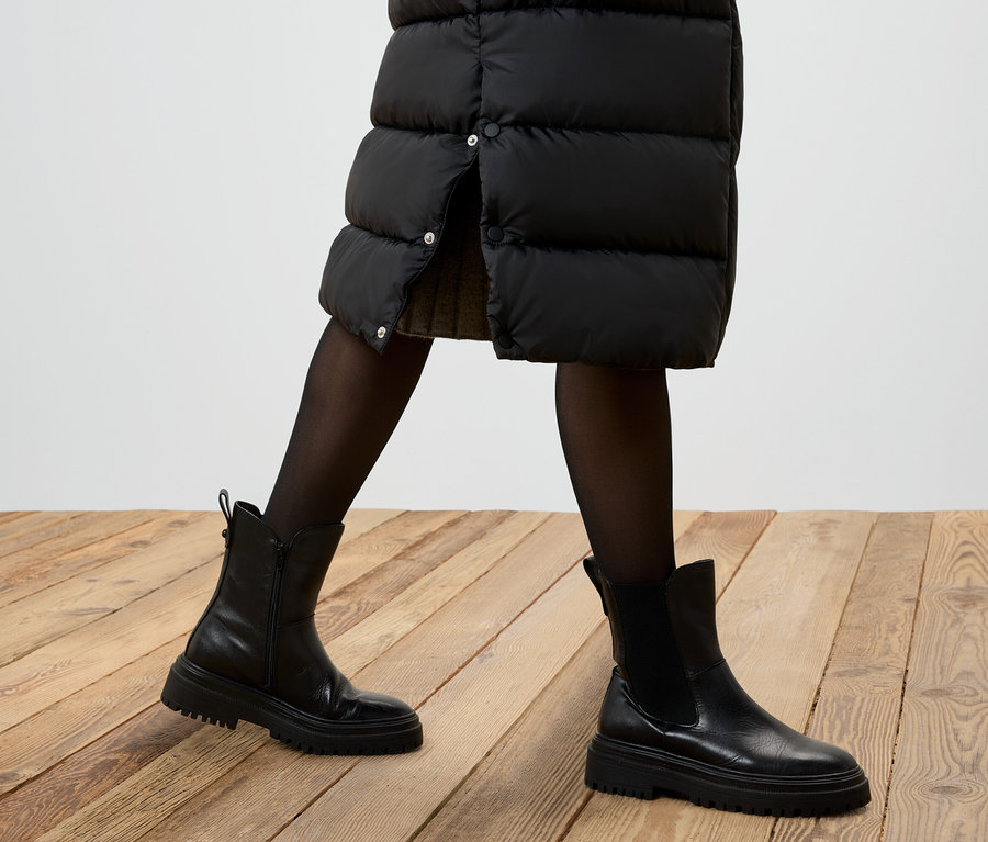 Čiastočný pohľad na osobu s čiernou páperovou sukňou, čiernymi pančuchami a čiernymi koženými topánkami na drevenej podlahe.