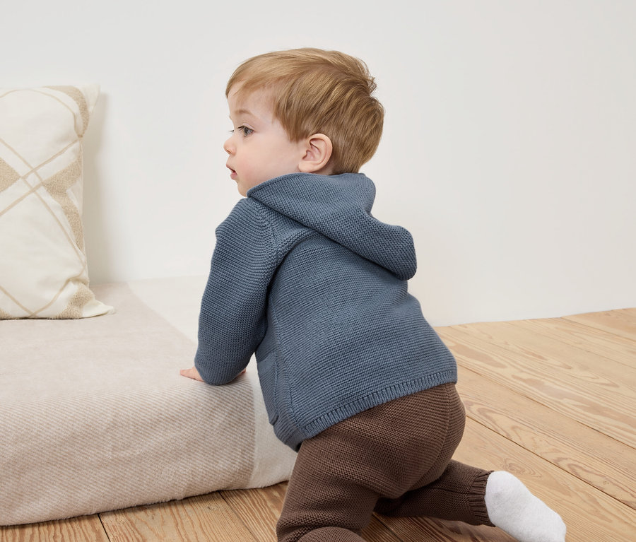 Dieťa lezie po béžovej pohovke v modrej mikine s kapucňou a hnedých nohaviciach.