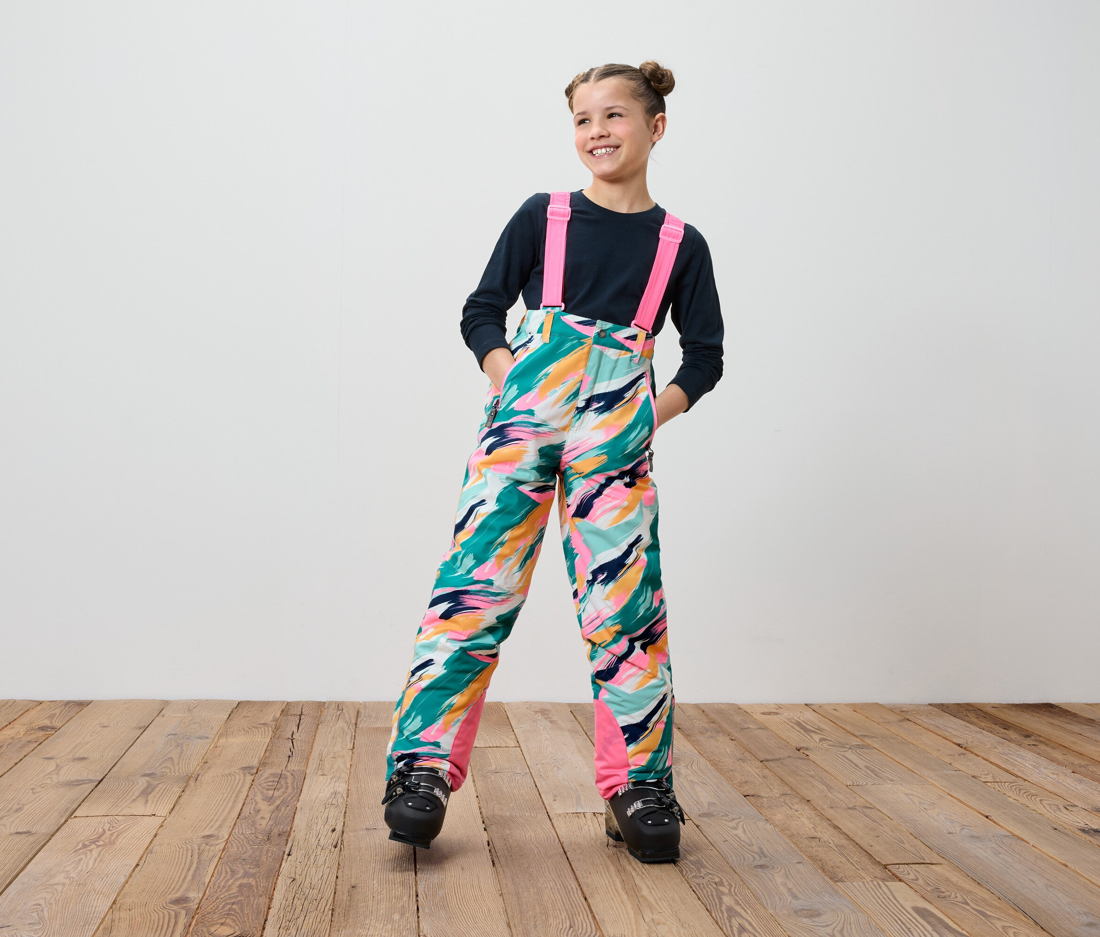 Usmievajúca sa dospievajúce dievča v farebných lyžiarskych nohaviciach s trakmi a čiernych lyžiarskych topánkach stojí na drevenej podlahe.