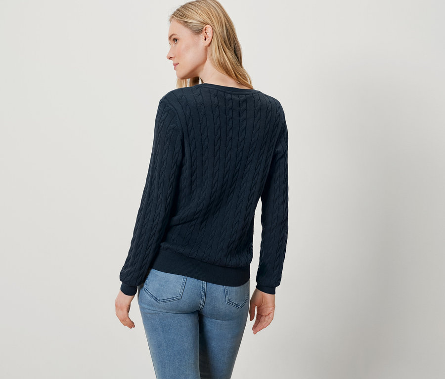 Žena s blond vlasmi, oblečená v modrom svetri a džínsoch, stojí chrbtom k fotoaparátu.