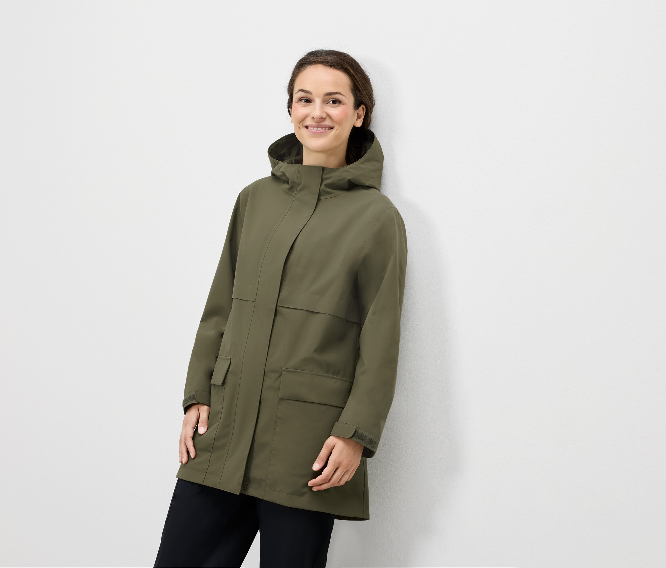 Žena pózuje v olivovozelenej bunde s kapucňou a čiernych nohaviciach.