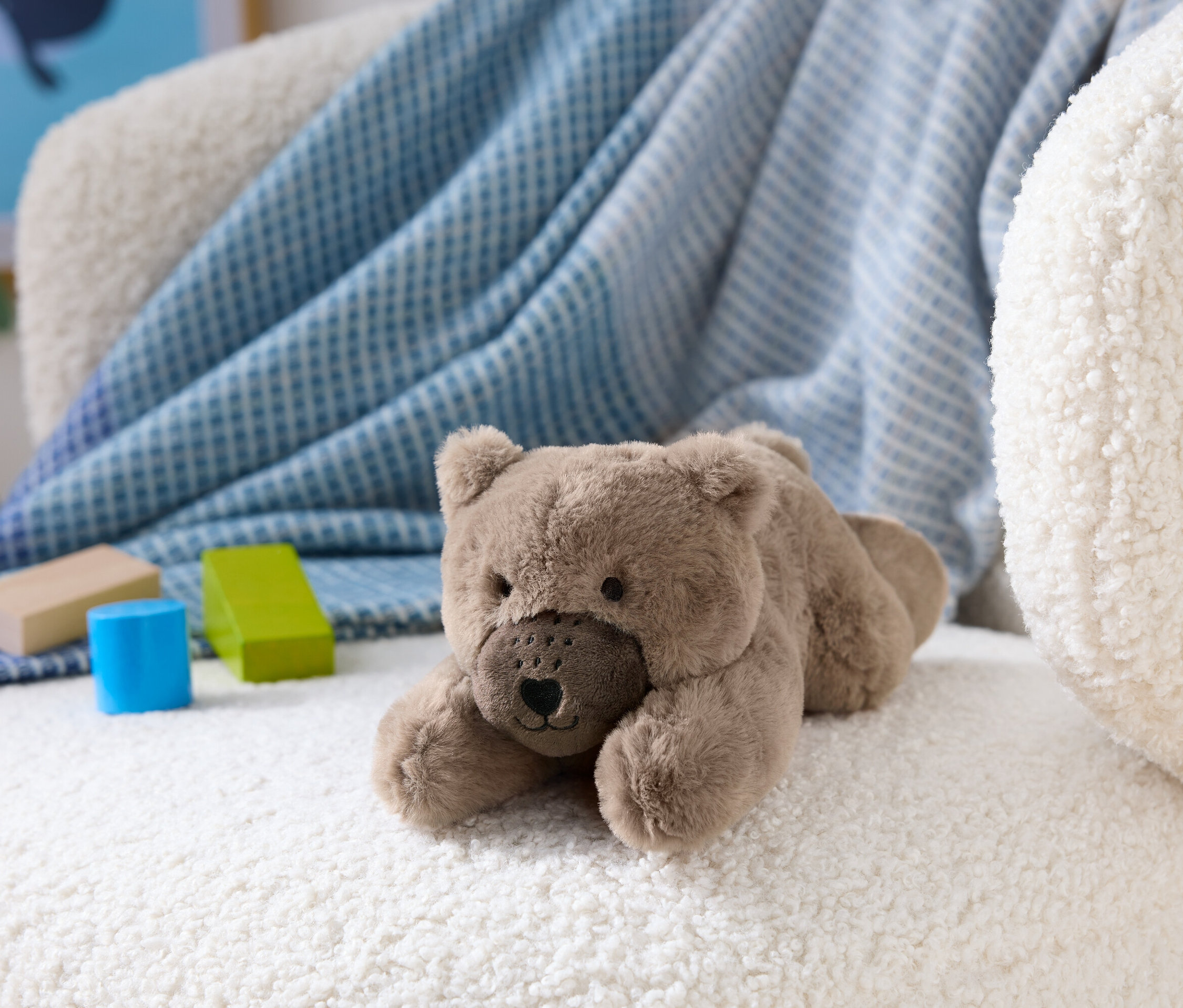 Hnedý plyšový medveď leží na bielom huňatom kresle vedľa drevených kociek.