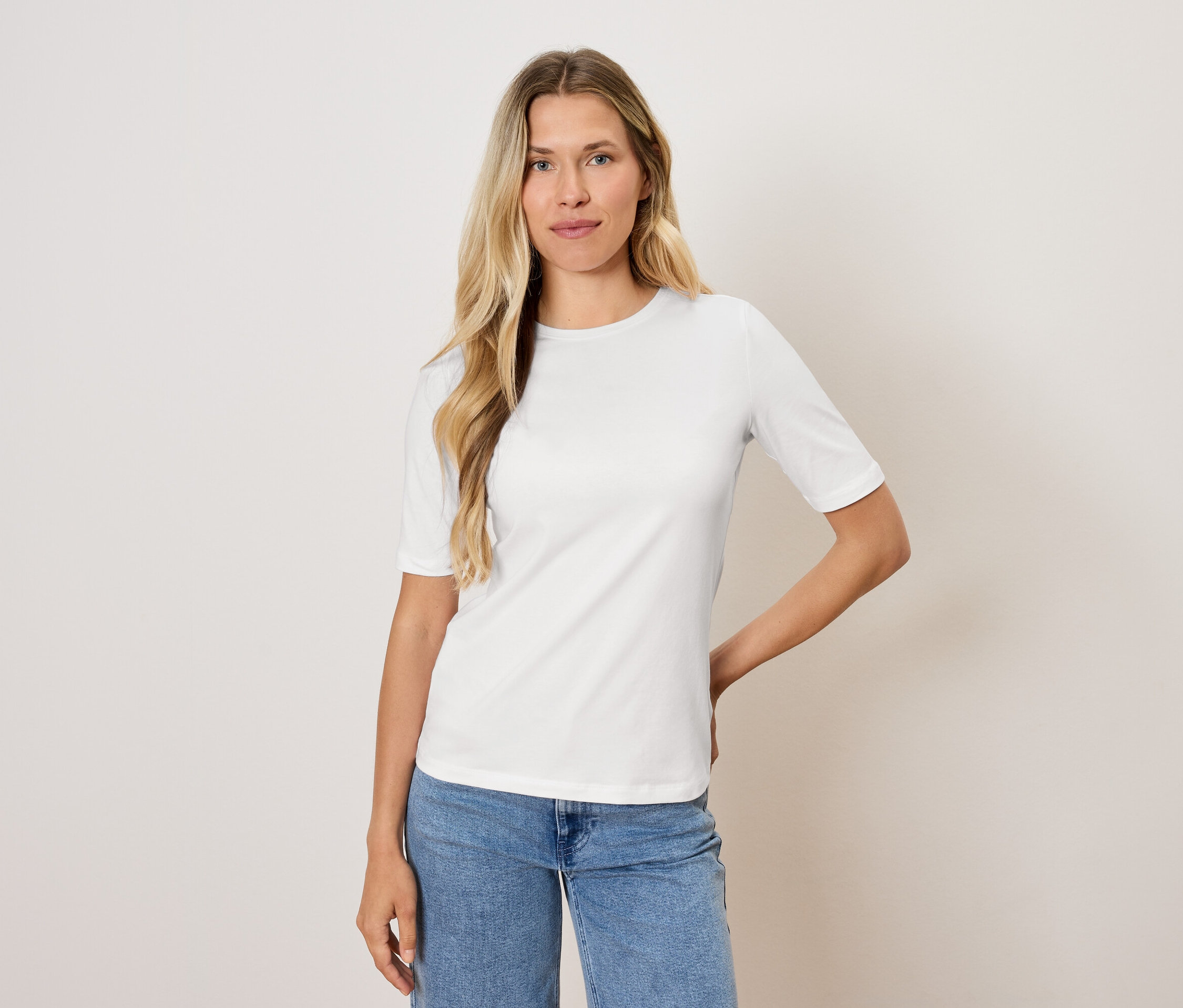 Žena s blond vlasmi pózuje v bielom tričku a modrých džínsoch.