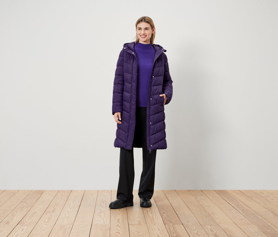 Žena stojí na drevenej podlahe a má na sebe fialový kabát, fialový sveter a čierne nohavice.