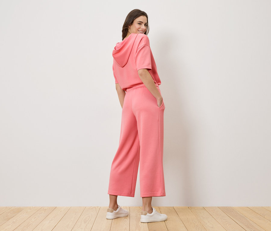 Žena s pohľadom cez plece má na sebe ružovú mikinu s kapucňou a culottes nohavice.