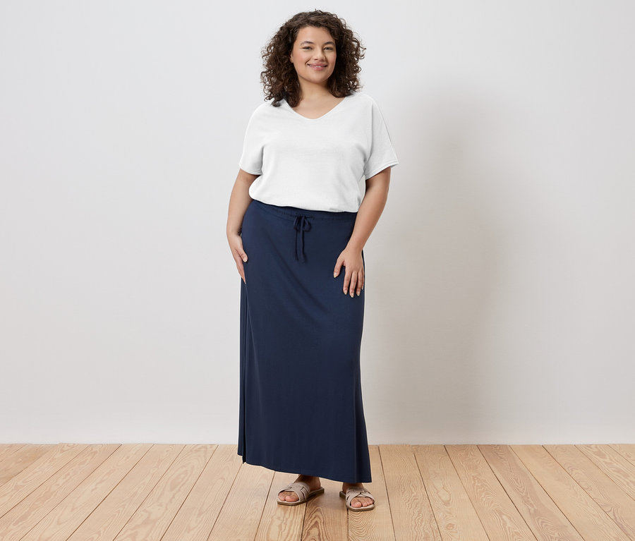 Žena stojí a má na sebe biele tričko, dlhú, námornícku modrú sukňu a sandále.