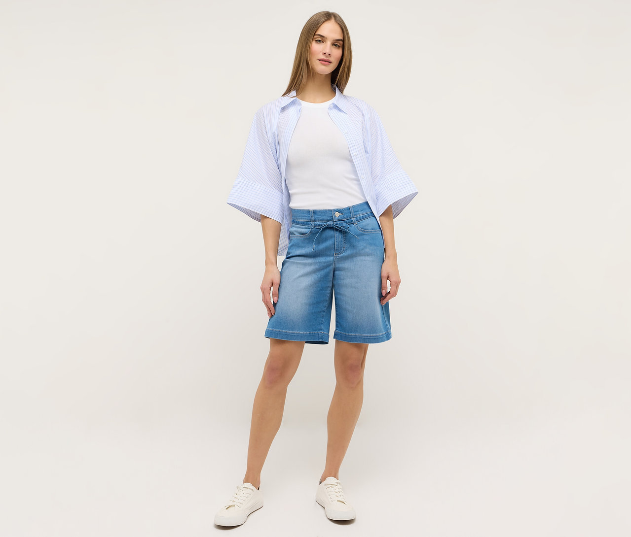 Žena má na sebe svetlomodré šortky Angels Denim »Lia City«, biele tričko, modrú pruhovanú blúzku a biele tenisky.