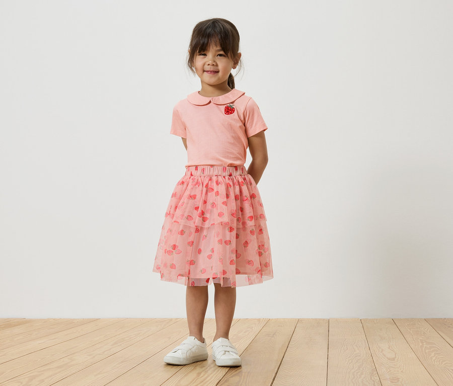 Dievča stojí pred bielou stenou a má na sebe ružové tričko s motívom jahody, ružovú tylovú sukňu s jahodami a biele tenisky.