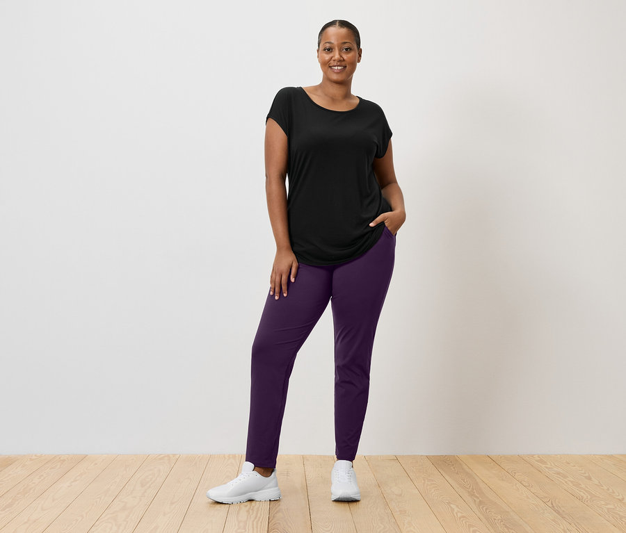 Žena v čiernom tričku, fialových nohaviciach a bielych teniskách stojí pred bielou stenou.