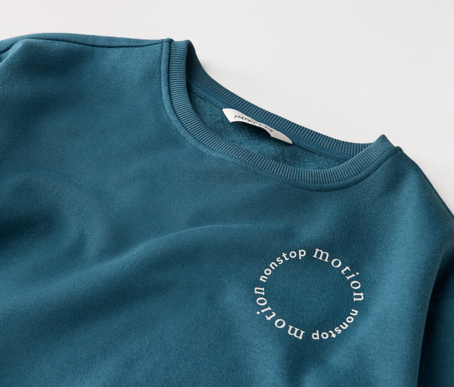 Detailný záber na petrolejovo modrú mikinu s nápisom "nonstop motion" v kruhu.