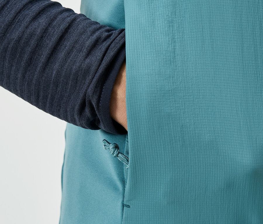 Detailný záber osoby, ktorá vkladá ruku do vrecka tyrkysovej bundy, s viditeľným námorníckym rukávom.